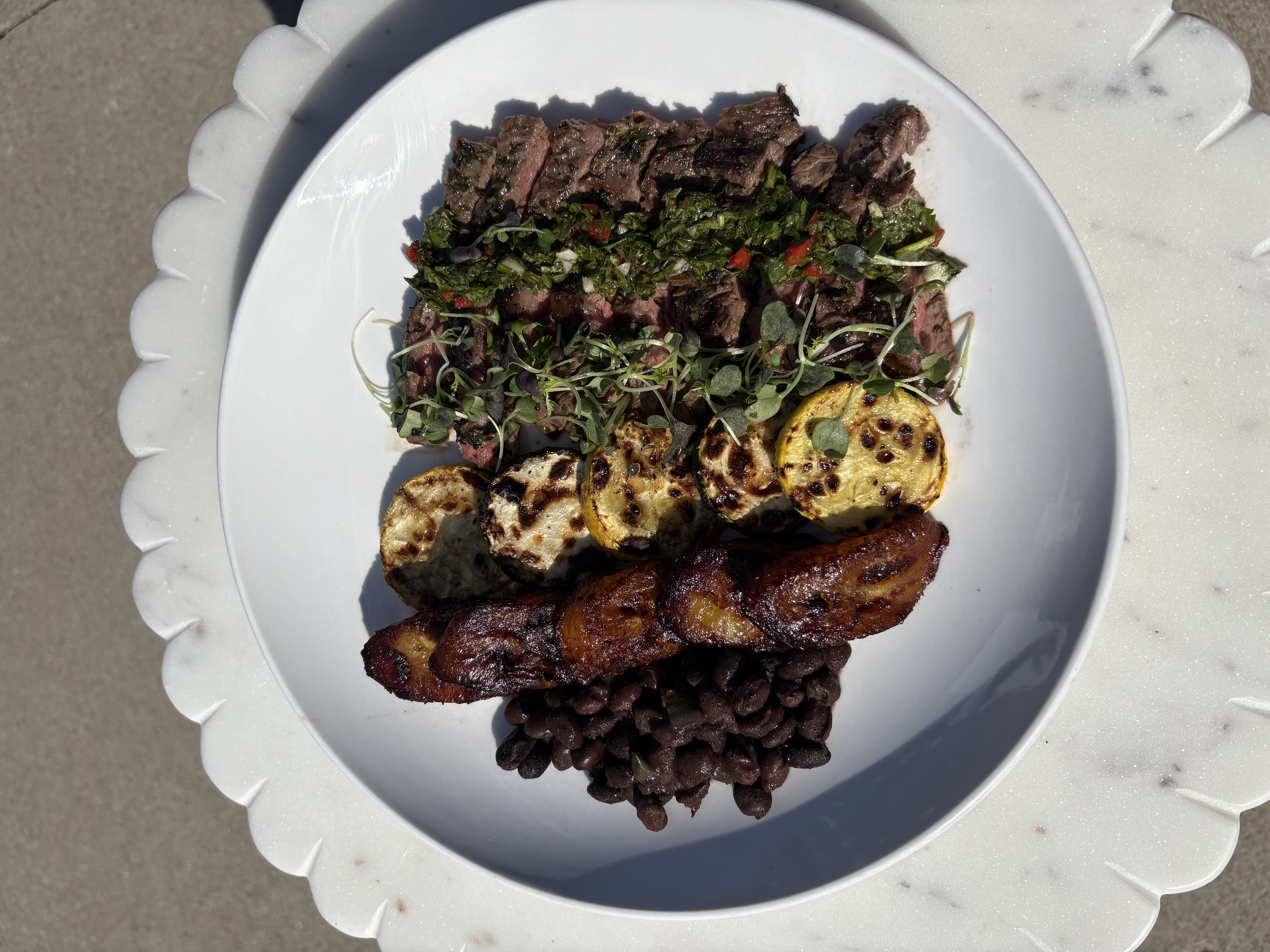 Chimichurri skirt steak plated on white scalloped plate with black beans, sweet plantains, and grilled zucchini